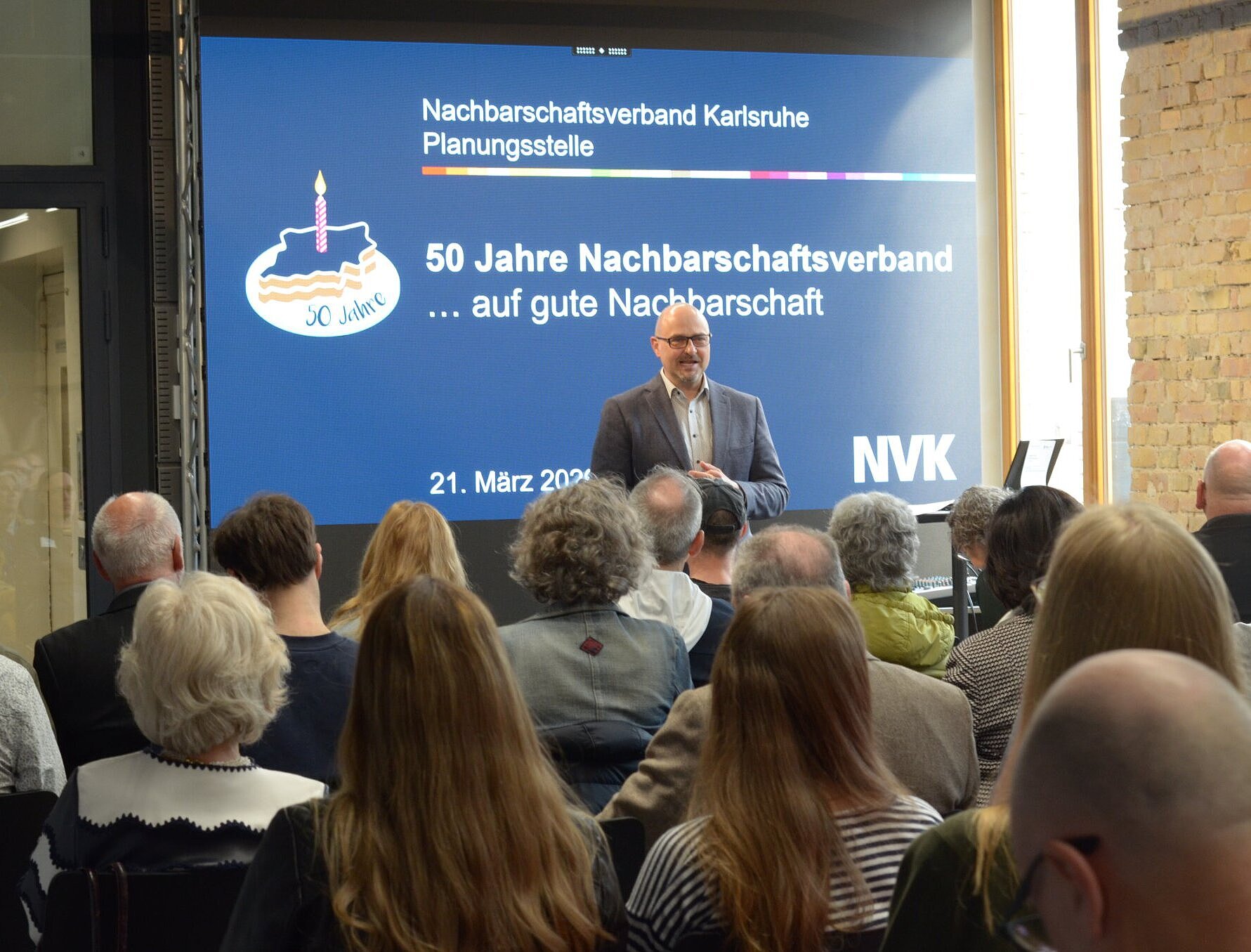 Oberbürgermeister  Johannes Arnold, als Verbandsvorsitzender hält eine Begrüßungsrede vor Publikum. Im Hintergrund Leinwand mit Begrüßungstext.